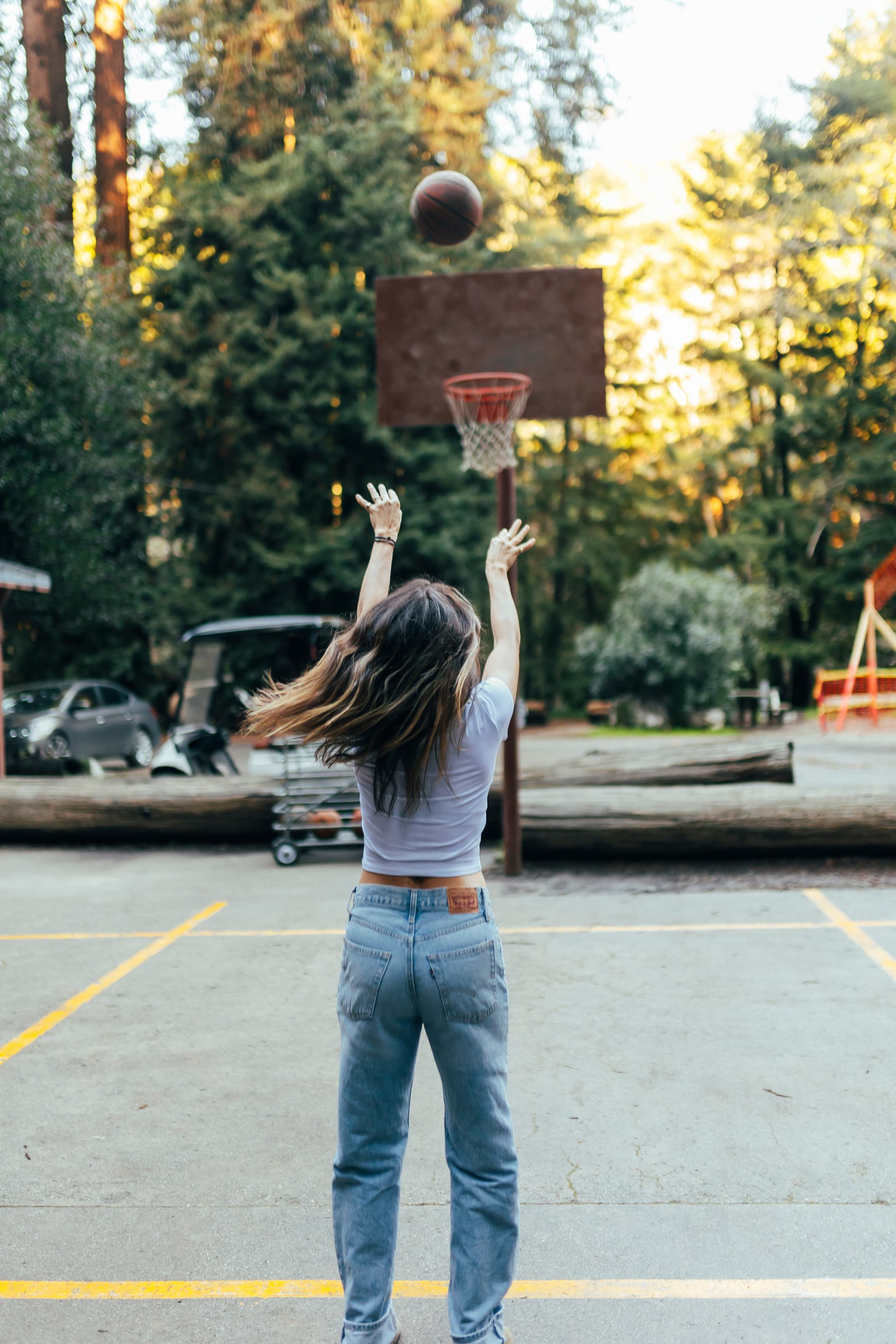 A young woman shooting a basketball toward a hoop outdoors in a parking lot, surrounded by trees at sunset.