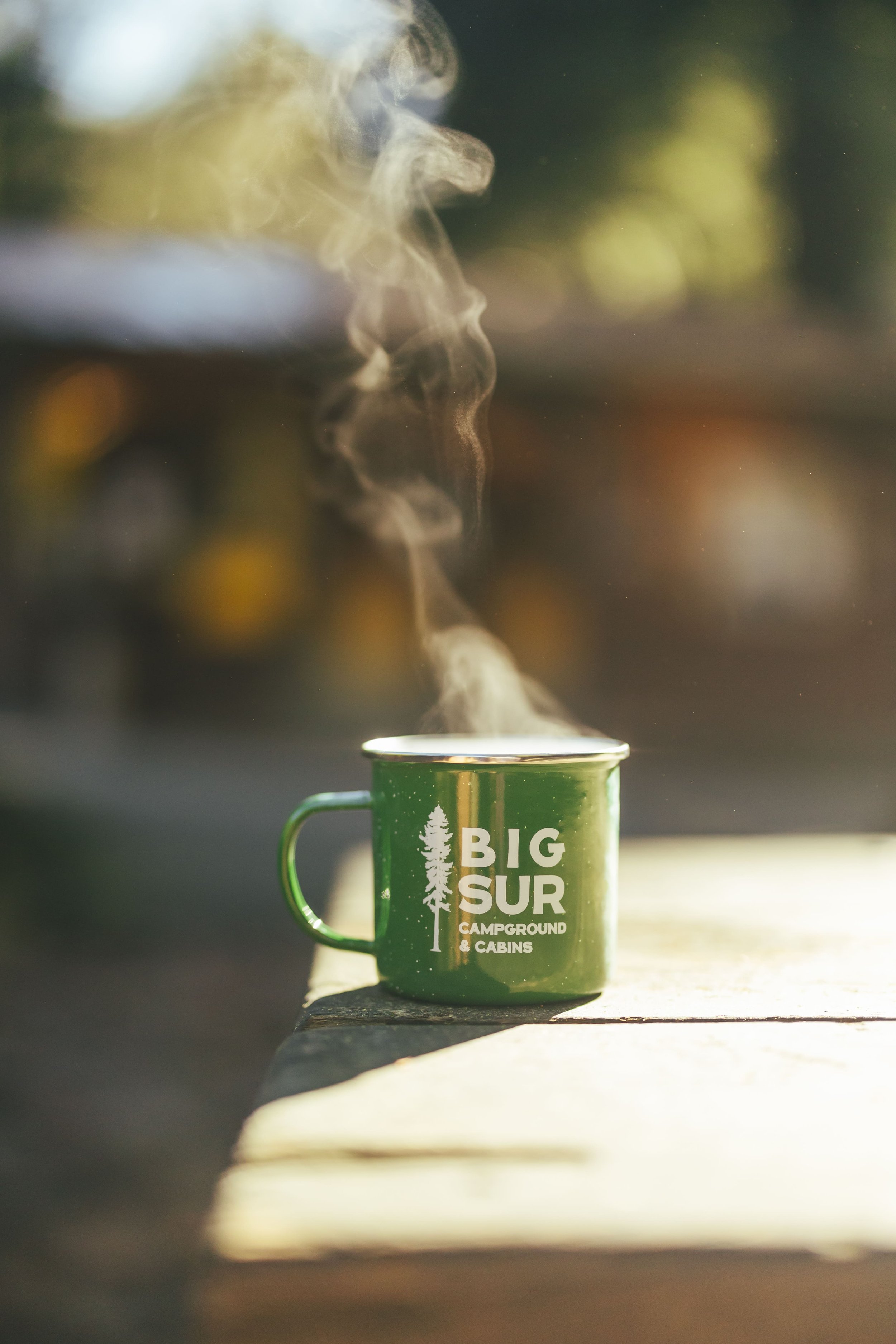 A green enamel camping mug with 'Big Sur Campground & Cabins' printed on it, placed on a wooden surface, with hot steam rising from it, outdoors with blurred trees in the background.